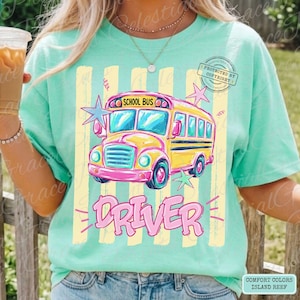 May include: A mint green t-shirt featuring a graphic of a yellow school bus with the text "SCHOOL BUS" above it and "DRIVER" below it. The design includes light yellow vertical stripes and pink and blue star accents on the front of the shirt.