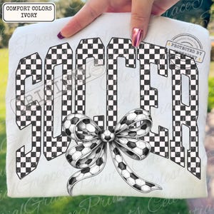 May include: Ivory-colored t-shirt with the word "SOCCER" in a checkered pattern. A soccer ball bow is centered below the word. The shirt is a Comfort Colors brand.