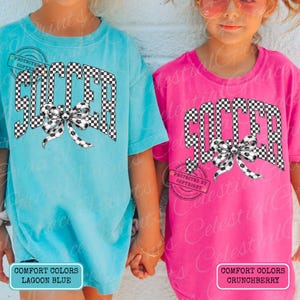 May include: Two short-sleeved t-shirts, one lagoon blue and one crunchberry pink, feature the word "SOCCER" in a checkered pattern with a soccer ball bow. The shirts have the text "COMFORT COLORS" and "PROTECTED BY COPYRIGHT" on them.