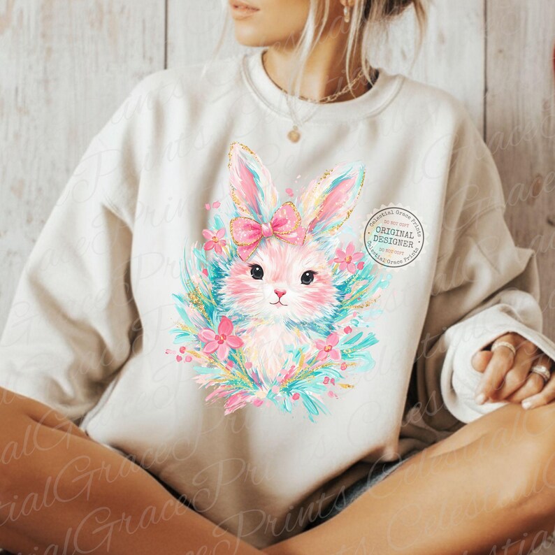 May include: A cream-colored sweatshirt featuring a watercolor illustration of a bunny with pink ears, a bow, and floral accents. The bunny is surrounded by a wreath of blue and pink flowers. The design includes the text "ORIGINAL DESIGNER".