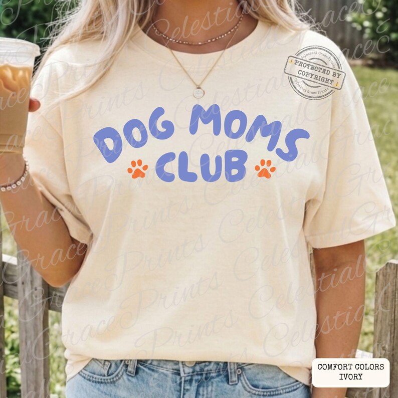 May include: Ivory-colored t-shirt with the text "DOG MOMS CLUB" in blue, with orange paw prints. The shirt is a Comfort Colors brand. The image also has a copyright notice.