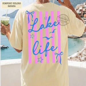 Puede incluir: Una camiseta amarilla clara con las palabras "Lake Life" en escritura azul, acentuada por rayas verticales rosas, lazos azules y estrellas. La camiseta tambi&eacute;n presenta im&aacute;genes de gaviotas. La esquina superior izquierda de la camiseta tiene las palabras "Comfort Colors Banana".