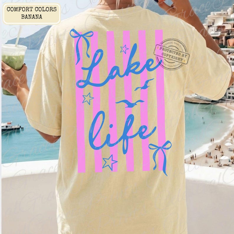 Puede incluir: Una camiseta amarilla clara con las palabras "Lake Life" en escritura azul, acentuada por rayas verticales rosas, lazos azules y estrellas. La camiseta tambi&eacute;n presenta im&aacute;genes de gaviotas. La esquina superior izquierda de la camiseta tiene las palabras "Comfort Colors Banana".
