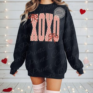May include: Black sweatshirt with the letters XOXO in a red and white checkered pattern. The letters are outlined in a tan color and have red bow accents. The sweatshirt has a crew neck and long sleeves.