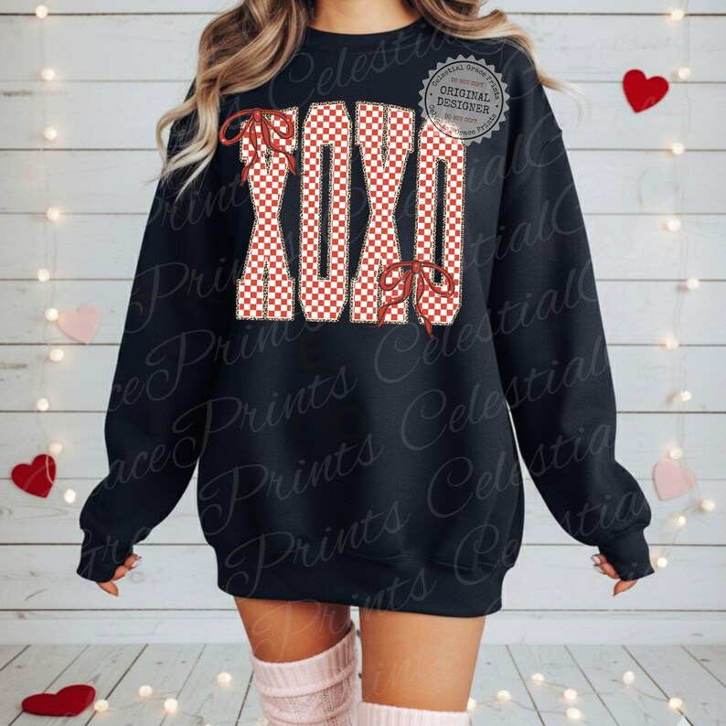 May include: Black sweatshirt with the letters XOXO in a red and white checkered pattern. The letters are outlined in a tan color and have red bow accents. The sweatshirt has a crew neck and long sleeves.