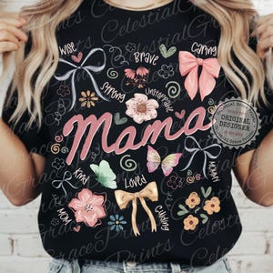 May include: Black t-shirt with the word "Mama" in pink script, surrounded by floral and bow graphics. Additional words like "Wise," "Brave," and "Caring" are also featured. The shirt has a round neckline.