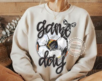 Könnte beinhalten: Cremefarbenes Sweatshirt mit einem Fußball-Grafikdesign und den Worten "Game Day" in schwarzer Schrift. Der Fußball hat schwarze, weiße, gelbe und orangefarbene Details. Über dem Ball befindet sich eine schwarze Schleife. Das Design ist auf dem Shirt zentriert.