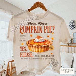 May include: An ivory t-shirt featuring a graphic of a pumpkin pie with whipped cream, surrounded by orange autumn leaves. Text reads: Farm Fresh PUMPKIN PIE? YES, PLEASE. EST. 1962, along with additional descriptive phrases about ingredients and baking.
