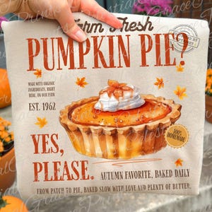 May include: A beige t-shirt featuring a graphic of a pumpkin pie with whipped cream, surrounded by orange maple leaves. Text reads: Farm Fresh PUMPKIN PIE? MADE WITH ORGANIC INGREDIENTS, RIGHT HERE, ON OUR FARM EST. 1962 YES, PLEASE. 100% HOMEMADE.