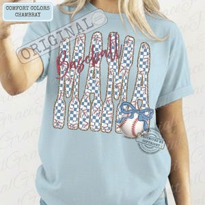 May include: Light blue chambray t-shirt with the text "Baseball Mama" in a stylized design. The letters are filled with a blue and white checkered pattern and baseball graphics. A baseball with a blue bow is featured.