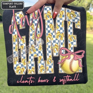 May include: Black t-shirt with the words "Day Game" in pink script. The letters are filled with a blue and white checkered pattern, yellow bows, and leopard print. A softball with a pink bow is featured below the text. The words "cleats, bows & softball" are written in pink script.