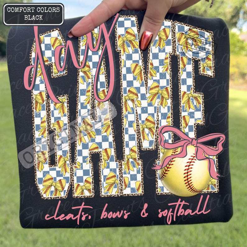 May include: Black t-shirt with the words "Day Game" in pink script. The letters are filled with a blue and white checkered pattern, yellow bows, and leopard print. A softball with a pink bow is featured below the text. The words "cleats, bows & softball" are written in pink script.