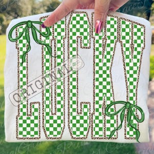 May include: White t-shirt with the word "JULY" in a green and white checkered pattern, outlined with a leopard print. Green bows are on the top left and bottom right. The word "ORIGINAL" is stamped across the shirt.