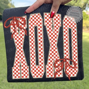 May include: Black sweatshirt featuring the letters "XOXO" in a red and white checkered pattern. The letters are outlined with a leopard print design and adorned with red bows. The sweatshirt is held by a hand with red nail polish.