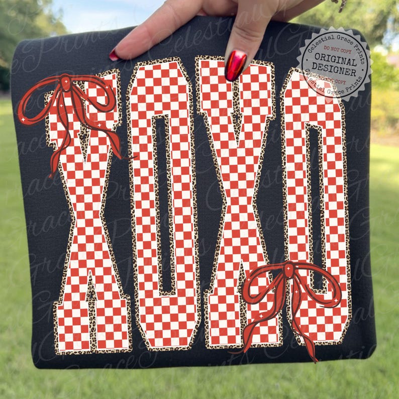 May include: Black sweatshirt featuring the letters "XOXO" in a red and white checkered pattern. The letters are outlined with a leopard print design and adorned with red bows. The sweatshirt is held by a hand with red nail polish.