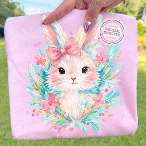 May include: A light pink sweatshirt featuring a detailed illustration of a bunny with a pink bow, surrounded by flowers and teal accents. The bunny has large black eyes and a pink nose. The text "Celestial Grace Prints" is visible.