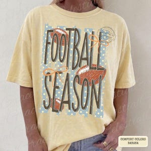 May include: A pale yellow short-sleeve t-shirt featuring the text FOOTBALL SEASON in large dark brown letters. The design includes illustrations of three brown footballs and orange line art bows over a light blue rectangular background with white dots.