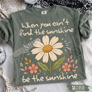 May include: A sage green crewneck t-shirt featuring a large white daisy graphic with a yellow center, surrounded by smaller pink flowers and green leaves. The text reads, "When you can't find the sunshine, be the sunshine" in a cream-colored font.