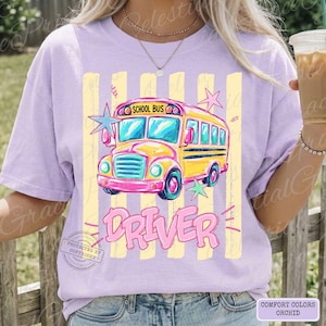 May include: A light purple t-shirt featuring a graphic of a yellow school bus with pink accents and the text "SCHOOL BUS" and "DRIVER" in pink lettering. The design includes light yellow vertical stripes and several light blue and pink stars.