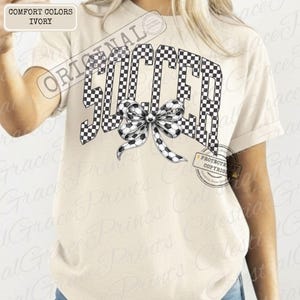 May include: Ivory-colored t-shirt with the word "SOCCER" in a black and white checkered pattern. A bow with soccer ball details is centered below the word. The shirt has short sleeves and a relaxed fit.
