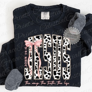 Puede incluir: Sudadera negra con la palabra "JESUS" en dise&ntilde;o de estampado de vaca, acentuada con un lazo rosa y el texto "John 14:6". La frase "the way, the truth, the life" est&aacute; impresa en la parte inferior. Se ve un calcet&iacute;n gris.