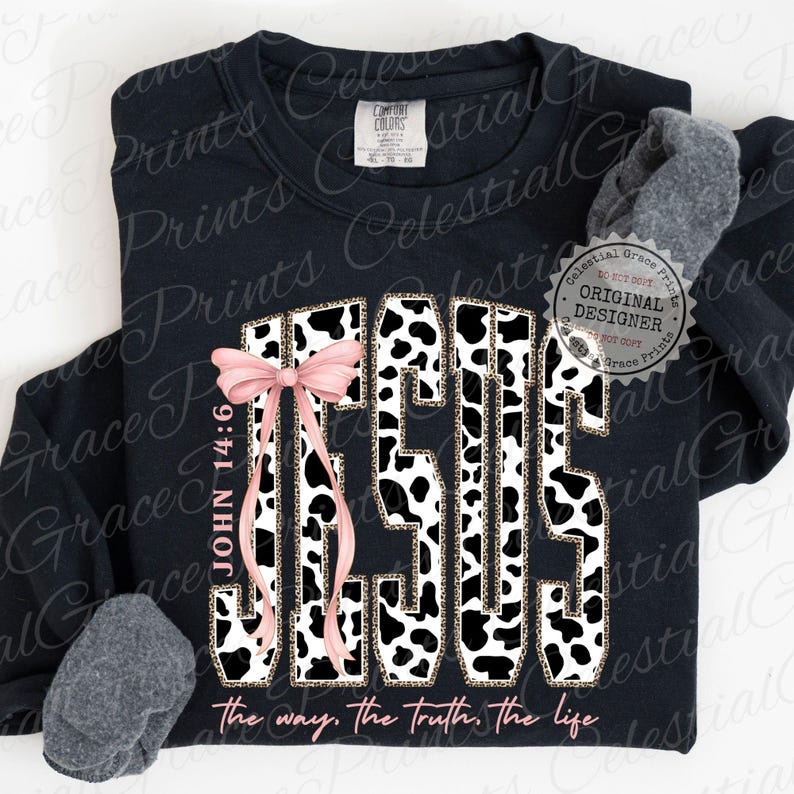 Puede incluir: Sudadera negra con la palabra "JESUS" en dise&ntilde;o de estampado de vaca, acentuada con un lazo rosa y el texto "John 14:6". La frase "the way, the truth, the life" est&aacute; impresa en la parte inferior. Se ve un calcet&iacute;n gris.