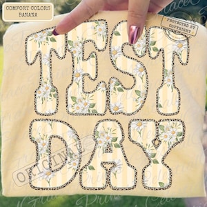 Peut inclure: Un t-shirt jaune avec l'inscription "TEST DAY" dans un motif floral. Les lettres sont soulign&eacute;es d'un motif l&eacute;opard. Le t-shirt porte une &eacute;tiquette de la marque Comfort Colors.