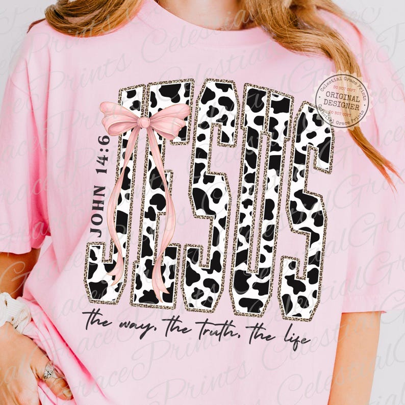Puede incluir: Camiseta rosa claro con un dise&ntilde;o gr&aacute;fico. La palabra "JESUS" est&aacute; en un estampado de vaca, adornada con un lazo rosa. El texto "JOHN 14:6" est&aacute; a la izquierda, y "the way, the truth, the life" est&aacute; en la parte inferior.
