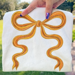 Op de afbeelding: Witte sweatshirt met een groot, gedetailleerd strikdesign in goud en oranje. De strik heeft een textuur met een glitterende omtrek, wat een feestelijke en elegante uitstraling creëert. Het ontwerp is gecentreerd op de voorkant van het kledingstuk.
