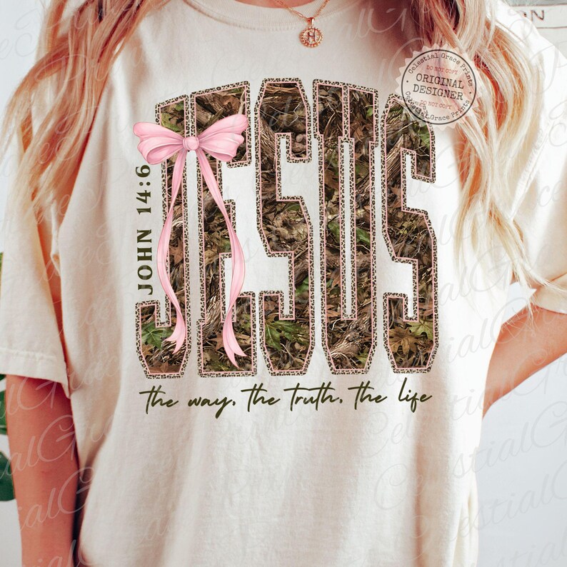 May include: Cream-colored t-shirt featuring the word "JESUS" in a camouflage and leopard print design, with a pink bow. The text "JOHN 14:6" is on the left side, and "the way. the truth. the life" is at the bottom.
