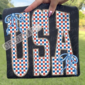 Op de afbeelding: Zwart T-shirt met het woord "USA" in een rood, wit en blauw geblokt patroon. De letters zijn omlijnd met een luipaardprint en versierd met blauwe strikken. Het shirt wordt door een hand omhoog gehouden.