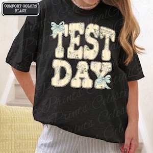 Peut inclure: T-shirt noir avec l'inscription "TEST DAY" en grandes lettres &agrave; motifs floraux. Les lettres sont jaunes avec des fleurs blanches et des n&oelig;uds bleu clair. Le t-shirt est noir.