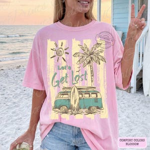 May include: Pink t-shirt with a beach-themed graphic. The design includes a sun, palm tree, surfboard, and a teal and white van with the text "Let's Get Lost". The shirt is a Comfort Colors Blossom.