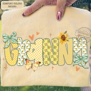 May include: A light yellow t-shirt with the word "GRANNY" in a playful font. The letters are decorated with sunflowers, gingham, and chevron patterns. The design includes bows, butterflies, and hearts.