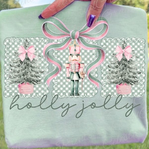 May include: A light green t-shirt with a festive Christmas design. The design features a nutcracker, Christmas trees, and a bow with pink and green ribbons. The words "holly jolly" are written in cursive below the design.