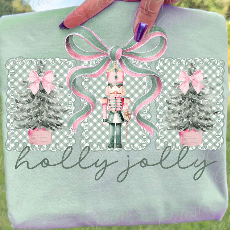 May include: A light green t-shirt with a festive Christmas design. The design features a nutcracker, Christmas trees, and a bow with pink and green ribbons. The words "holly jolly" are written in cursive below the design.
