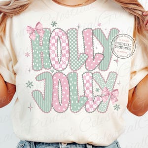 以下が含まれることがあります： クリーム色のTシャツに「HOLLY JOLLY」の文字が祝祭的なデザインで描かれています。文字はピンクとミントグリーンで、ギンガムチェックやストライプなどのパターンがあり、リボンと雪の結晶がアクセントになっています。