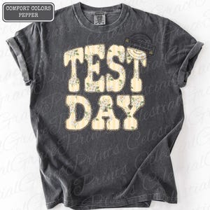 Peut inclure: T-shirt gris fonc&eacute; avec l'inscription "TEST DAY" dans un motif floral et ray&eacute;. Le texte est en gras, avec un motif floral jaune clair et blanc. Le t-shirt a des manches retrouss&eacute;es.