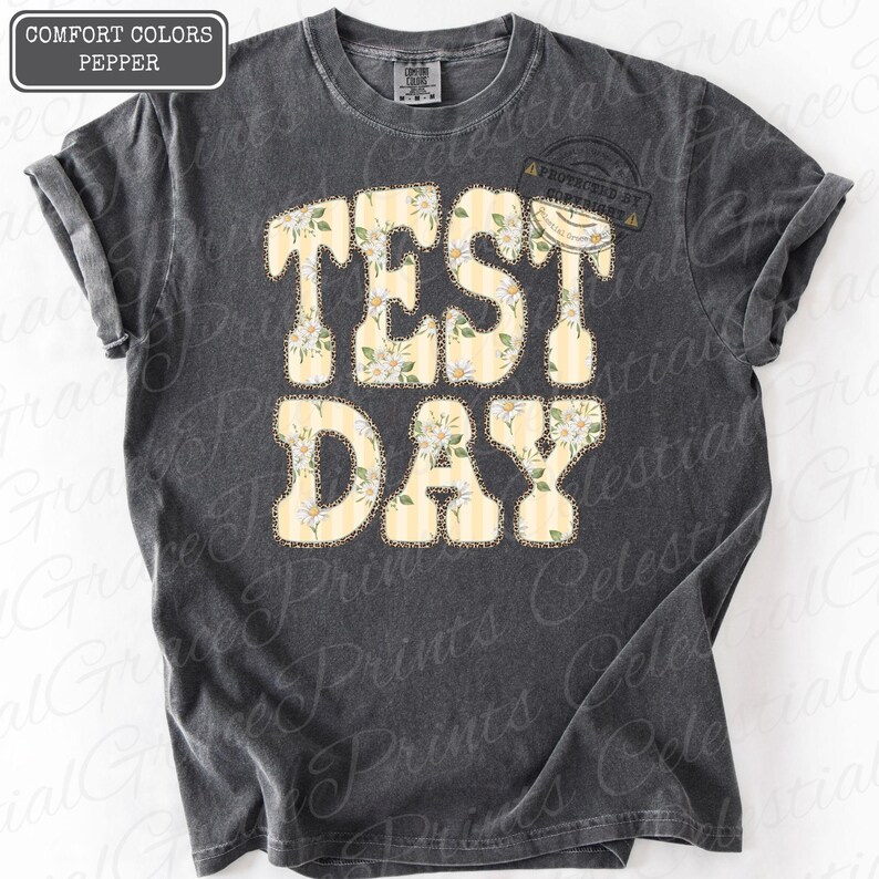 Peut inclure: T-shirt gris fonc&eacute; avec l'inscription "TEST DAY" dans un motif floral et ray&eacute;. Le texte est en gras, avec un motif floral jaune clair et blanc. Le t-shirt a des manches retrouss&eacute;es.
