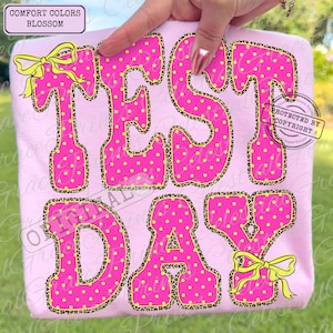 May include: Pink t-shirt with the words "TEST DAY" in large, bubble letters. The letters are hot pink with yellow polka dots and leopard print trim. Yellow bows accent the letters. The shirt is labeled "COMFORT COLORS BLOSSOM".