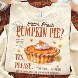 May include: A cream-colored crewneck sweatshirt featuring a graphic of a pumpkin pie with whipped cream. Text reads: Farm Fresh PUMPKIN PIE? EST. 1962, YES, PLEASE. AUTUMN FAVORITE, BAKED DAILY, MADE WITH ORGANIC INGREDIENTS, RIGHT HERE, ON OUR FARM.