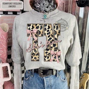 Può includere: Una maglietta grigia Comfort Colors con un motivo leopardato che forma il numero "74" con fiocchi rosa e le parole "She's from" in corsivo rosa. La maglietta è indossata con jeans blu e una cintura nera.