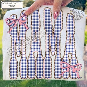 May include: Ivory t-shirt featuring the word "MAMA" in large letters. The letters are filled with a blue and white gingham pattern and outlined with a leopard print. Pink bows are placed on the top and bottom of the word.
