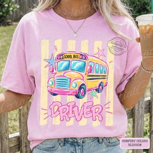 May include: A light pink short-sleeve t-shirt featuring a graphic of a yellow school bus with the text "SCHOOL BUS" above it and "DRIVER" below it. The design includes vertical pale yellow stripes, pink and teal stars, and a copyright stamp.