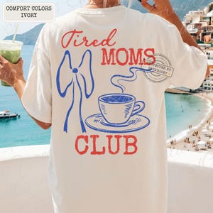 May include: Ivory-colored t-shirt with the words "Tired Moms Club" in red and blue. The design includes a blue bow and a blue coffee cup with steam. The shirt is a comfort color.