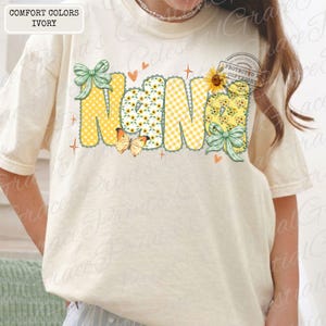 May include: Ivory-colored t-shirt with the word "Nana" in a whimsical design. The letters feature polka dots, sunflowers, and gingham patterns, accented with butterflies, bows, and hearts. The shirt is labeled "Comfort Colors Ivory."