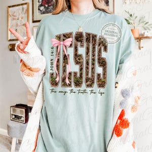 May include: A light blue t-shirt with the word "JESUS" in a camouflage pattern, accented with a pink bow. The text "JOHN 14:6" is on the left side, and "the way, the truth, the life" is at the bottom. A circular logo is on the upper right.
