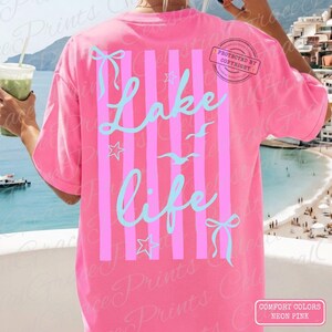 Puede incluir: Camiseta rosa ne&oacute;n con rayas rosas verticales y las palabras "Lake Life" en escritura azul claro. El dise&ntilde;o incluye estrellas, lazos y gaviotas. La camiseta es de la marca Comfort Colors.