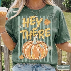 Puede incluir: Camiseta verde con las palabras "HEY THERE" en amarillo, un gráfico de calabaza y detalles de hojas de otoño. La camiseta tiene rayas verticales y una etiqueta "COMFORT COLORS MOSS".