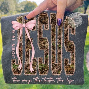 May include: Dark gray t-shirt with the word "JESUS" in a camouflage pattern outlined in leopard print. A pink bow is tied to the left side. The text "JOHN 14:6" and "the way, the truth, the life" are also visible.